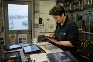 Woman in her forties learning new skills in a coastal Breton workshop, surrounded by tablets, tools, and training materials, with soft daylight and a harbor visible outside.