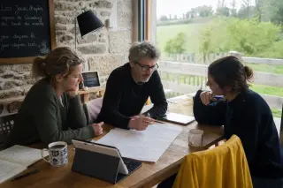 Three adults collaborating around a wooden table in a bright rural coworking space in Brittany, focused on handwritten notes and digital tablets, with countryside visible through large windows.
